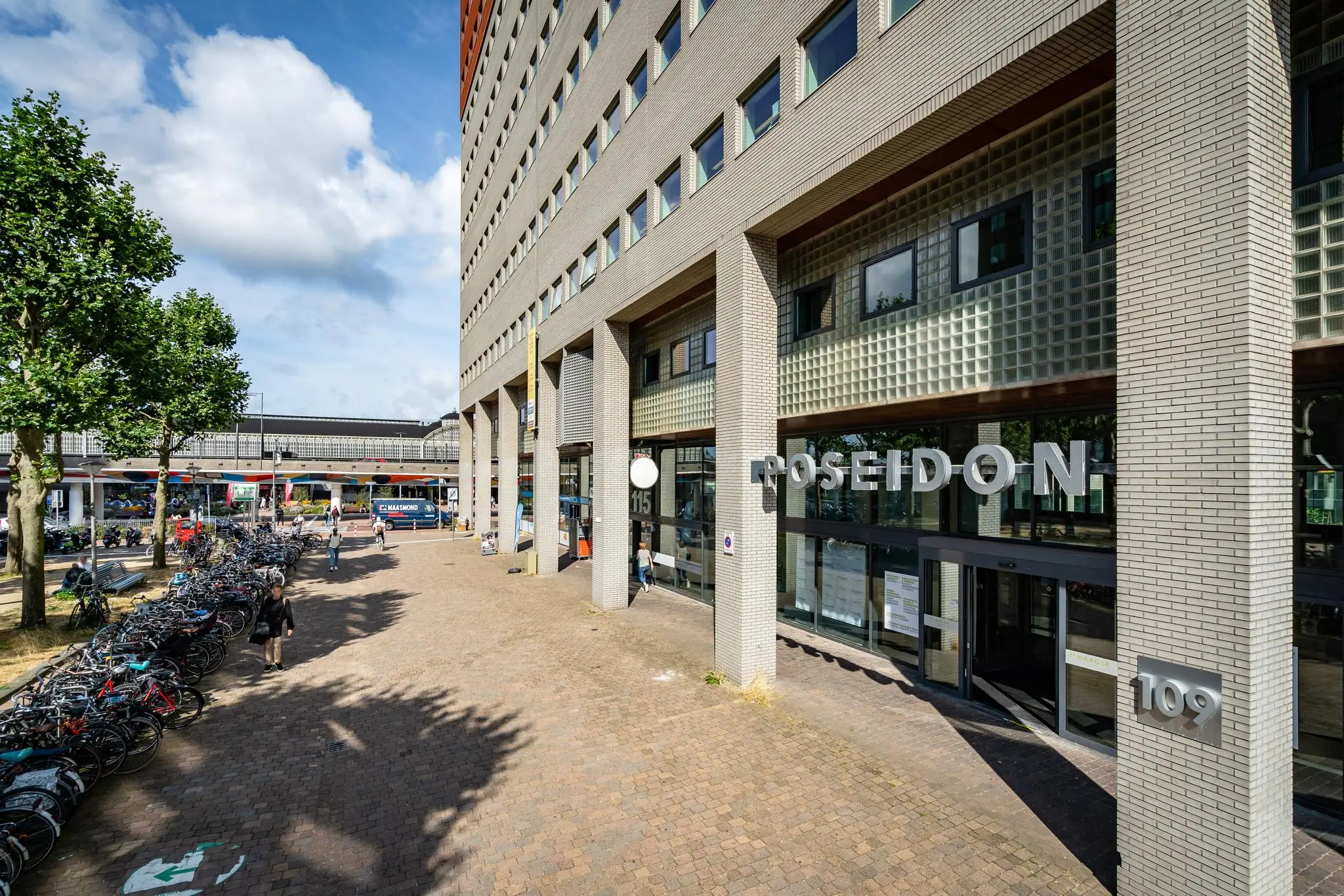 Entree van het Poseidon-gebouw aan het Johanna Westerdijkplein met fietsenstalling en voorbijgangers op de voorgrond.